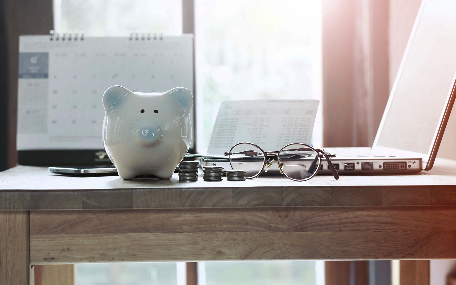 A piggy bank, glasses, laptop, calendar and loose change on top of a desk