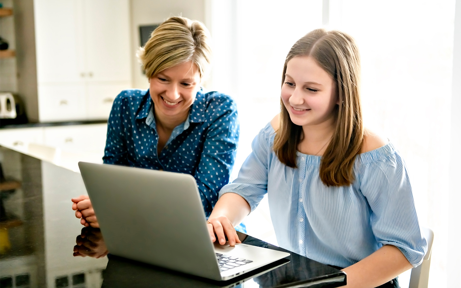 A mother using a laptop in kitchen with teenager