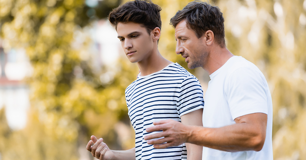 father gesturing and talking with teenager son in park 
