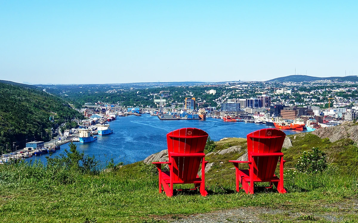 Two red chairs sitting on top of a hill over looking a bay full of boats with a city sprawling out into the background