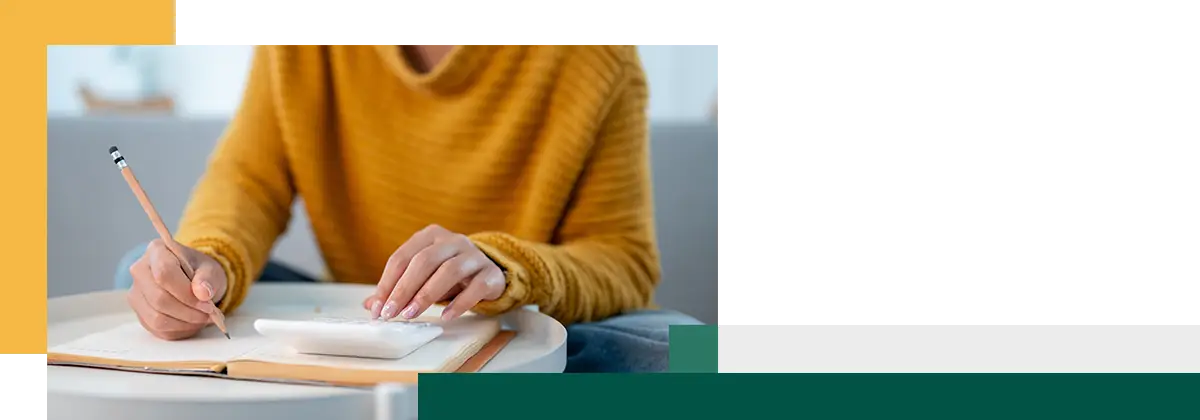 A closeup image of a woman sitting on a sofa at home writing in a book and using a calculator