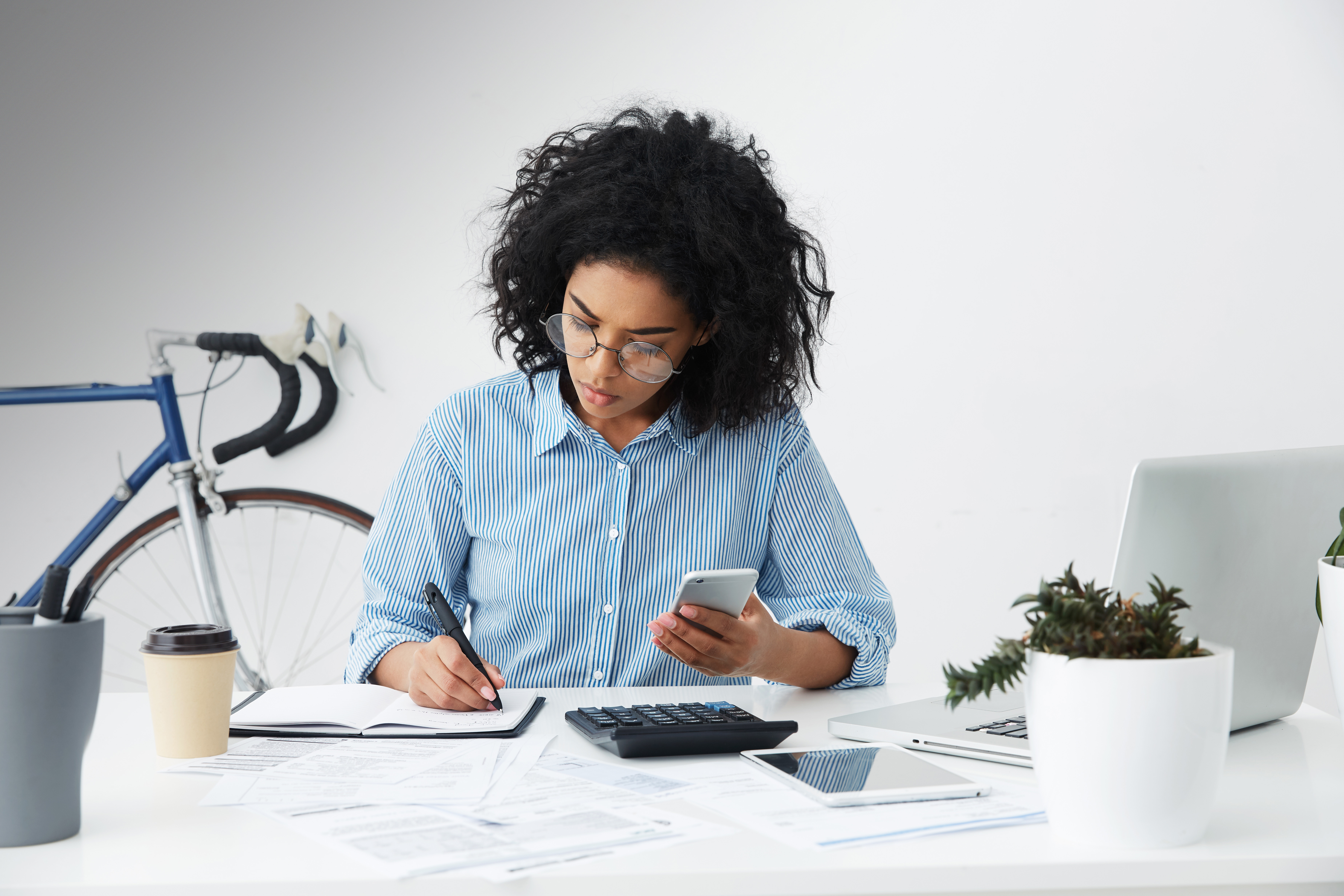 Concentrated female holding phone and making notes while planning budget and calculating bills