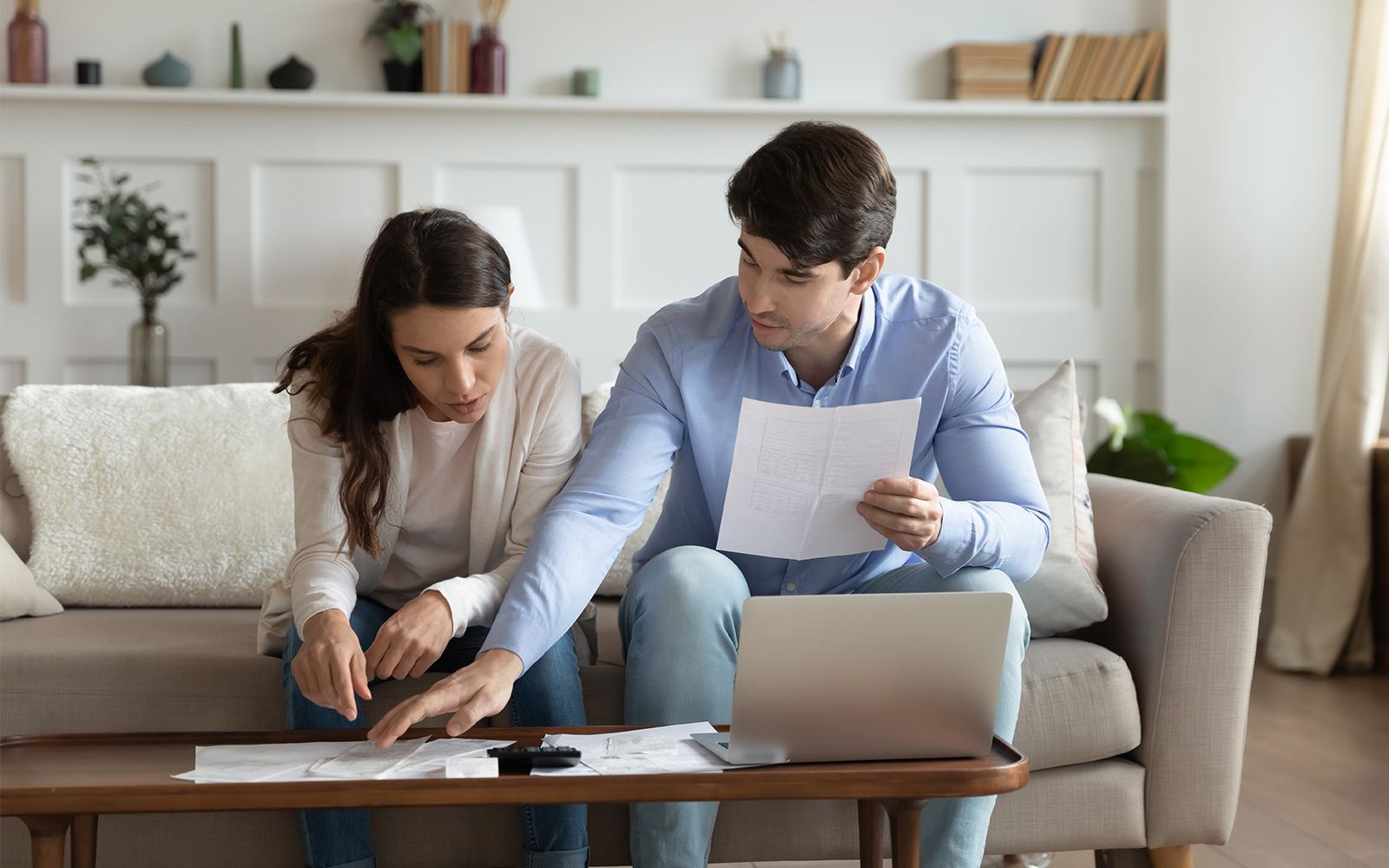 Concentrated spouses sitting on couch at home discussing with laptop and calculator