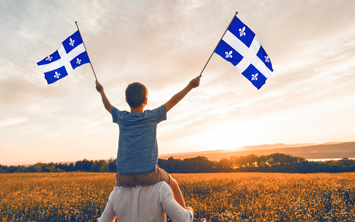 Child sitting on father's shoulders holding two Quebec flags.