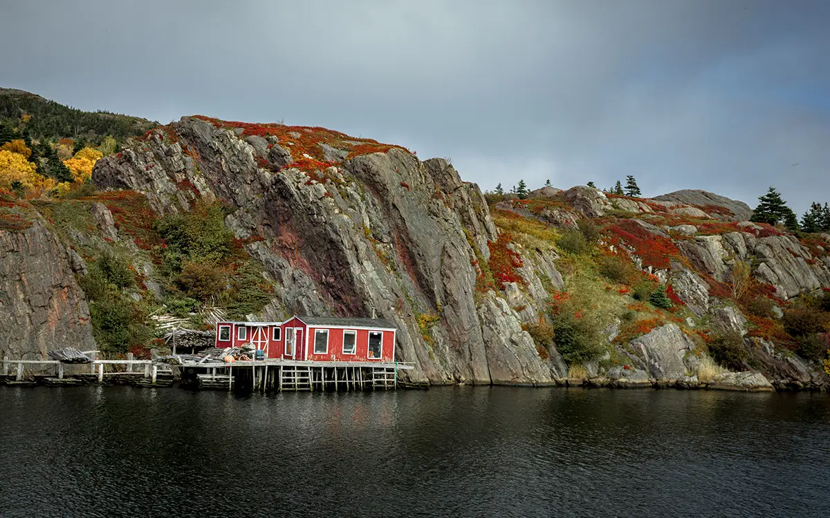 A red building sits on a wooden pier by the water, nestled against steep rocky cliffs covered in autumn foliage. The sky is overcast, and the vibrant fall colors of red, orange, and green stand out against the gray rock.