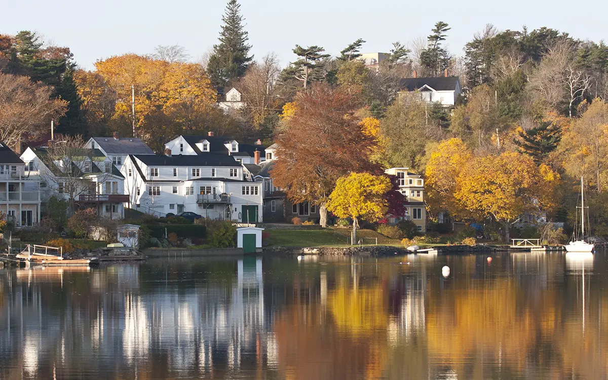 A serene lakeside community with white houses along the shoreline, surrounded by trees displaying vibrant yellow, orange, and red autumn leaves. The calm water reflects the houses and trees, creating a peaceful scene.