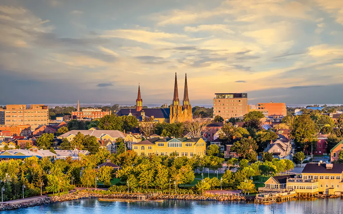 A scenic view of a town at sunset with historic buildings and churches featuring tall steeples. The foreground includes a park with green trees and a body of water, while the sky glows with warm colors as the day transitions into evening.