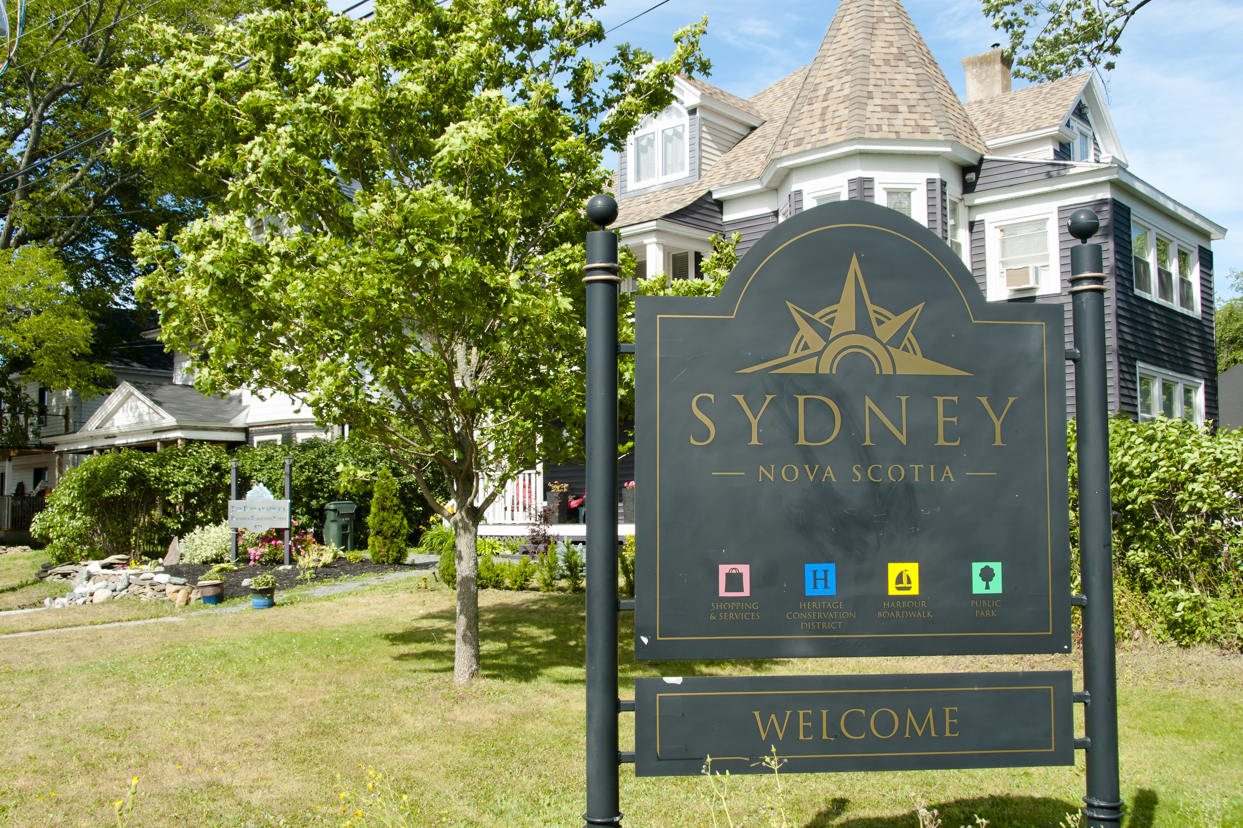 Sign in front of house that says "Sydney, Nova Scotia. Welcome. Shopping & Services; Heritage Conservation District; Harbour Boardwalk; Public Park."