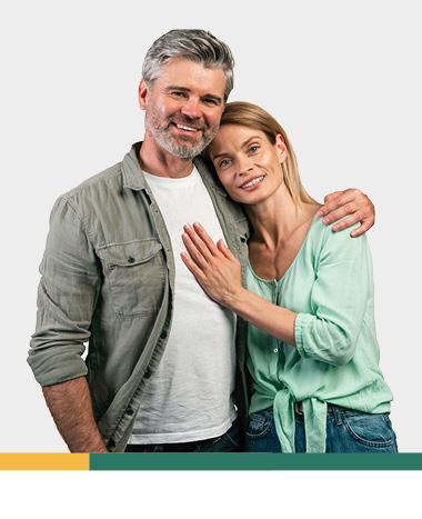 Smiling couple standing together, one person resting their head on the other’s shoulder, both wearing casual clothing against a light background.