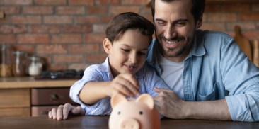 A man and a boy are at a table with a piggy bank, engaging in a discussion about saving and financial responsibility.