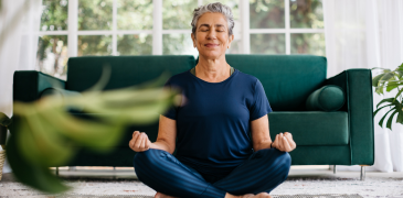 A person with short gray hair sits cross-legged on a floor, meditating with eyes closed in front of a green sofa and large window. A houseplant is blurred in the foreground.