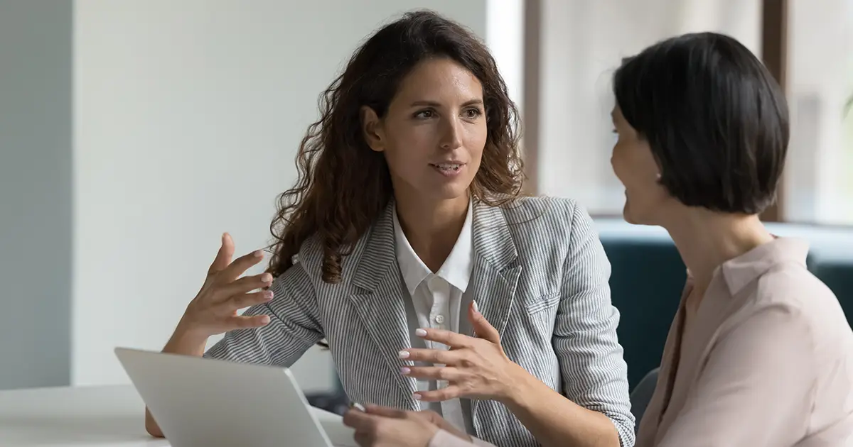 Two business women sit at desk discuss project details