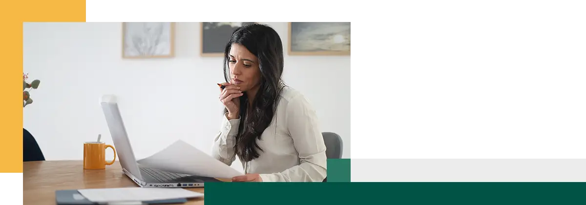 woman is working from home and looking over paperwork while biting on a pen