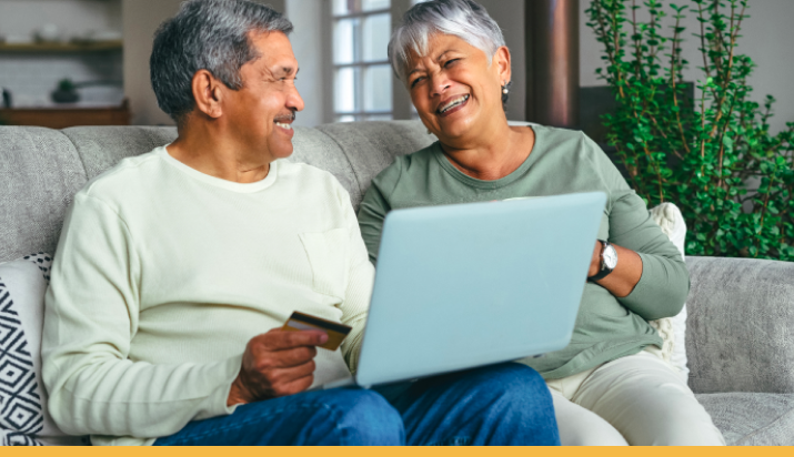 An elderly couple sitting on a couch, smiling and engaging with a laptop. The man holds a credit card, while the woman laughs, creating a warm and happy atmosphere. There are decorative plants in the background, adding to the cozy setting.
