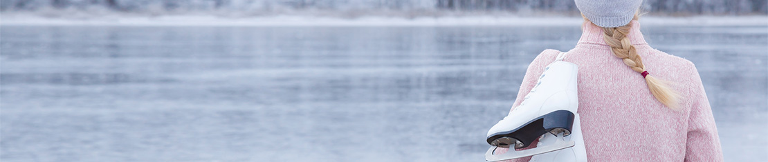 Woman wearing a pink coat and purple toque with a pair of skates over her shoulder standing in front of a frozen body of water